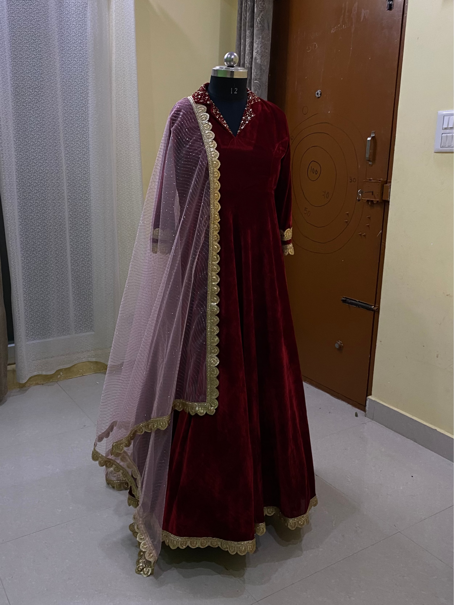 Red velvet dress with a sheer dupatta on a mannequin in a room with curtains and a door.